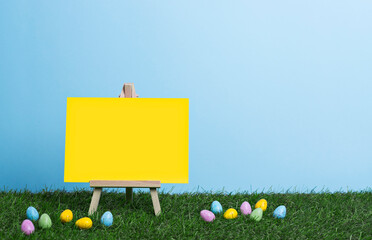 Blank yellow canvas on an easel, place for text, stands on the grass among scattered painted Easter...