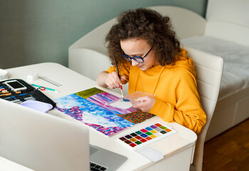 Caucasian teenage girl is drawing with paints sitting at the table. Online distance education concept.