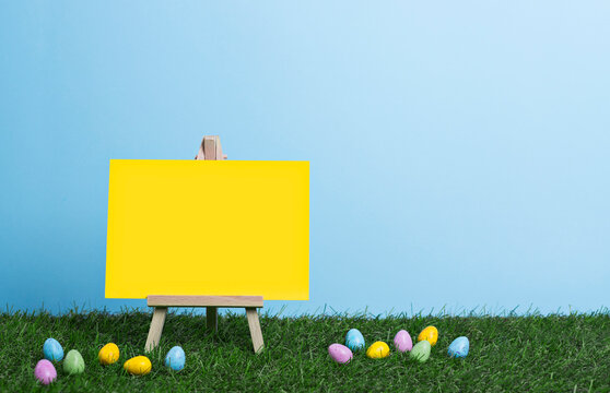Blank yellow canvas on an easel, place for text, stands on the grass among scattered painted Easter eggs on the background of clear blue sky, Easter banner
