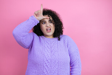 Young beautiful woman wearing casual sweater over isolated pink background making fun of people with fingers on forehead doing loser gesture mocking and insulting.