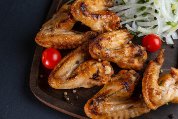grilled on coals chicken wings on a wooden board with pickled onions, chili peppers and cherry tomatoes on a dark background. photos with a small depth of field, top view