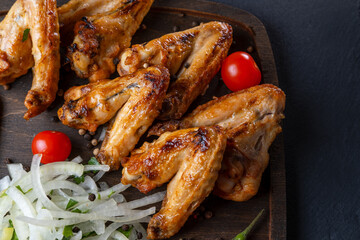 grilled on coals chicken wings on a wooden board with pickled onions, chili peppers and cherry tomatoes on a dark background. photos with a small depth of field, top view