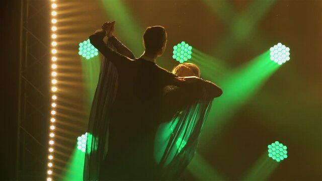 Silhouetted pair of ballroom dancers practicing dancing jumping holding hands. Classical dance choreography in dark studio against backdrop of multicolored spotlights. Close up. Slow motion.
