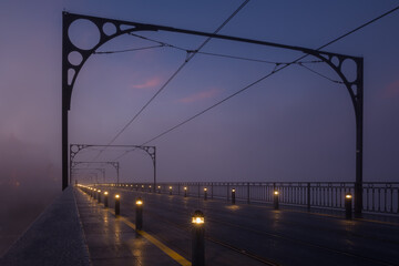 City street, a bridge with tram tracks. The foggy morning before dawn. Blue sky with pink clouds. Rows of low lanterns along the edges of the road. Linear perspective.