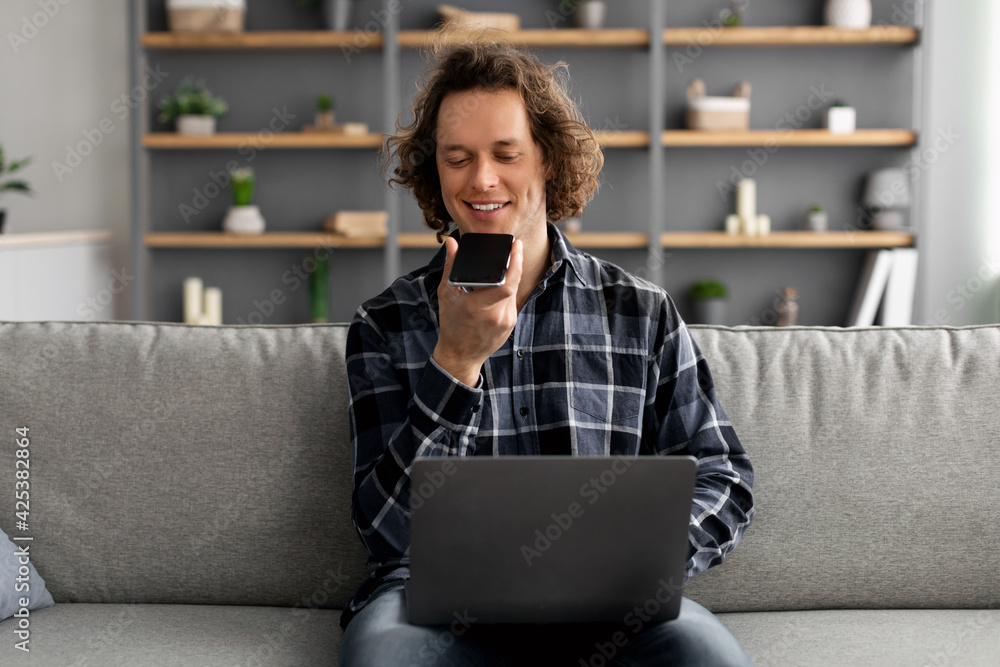 Poster man using voice assistant on smartphone working on laptop indoor