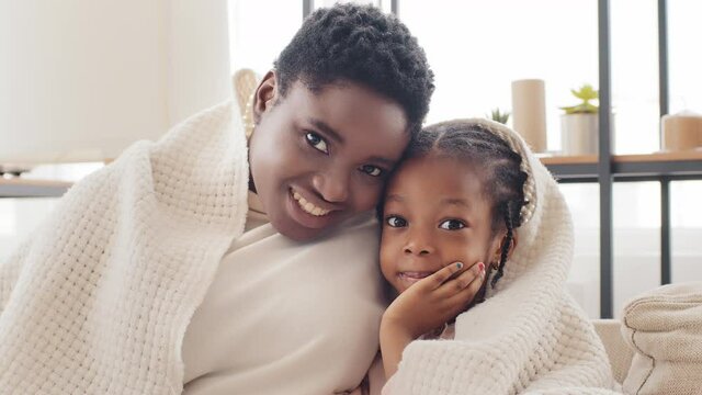 Portrait of afro american family at home covered by blanket, african single mother adult black woman hugs cuddles to little cute mixed race girl child kid daughter looking at camera with happy faces