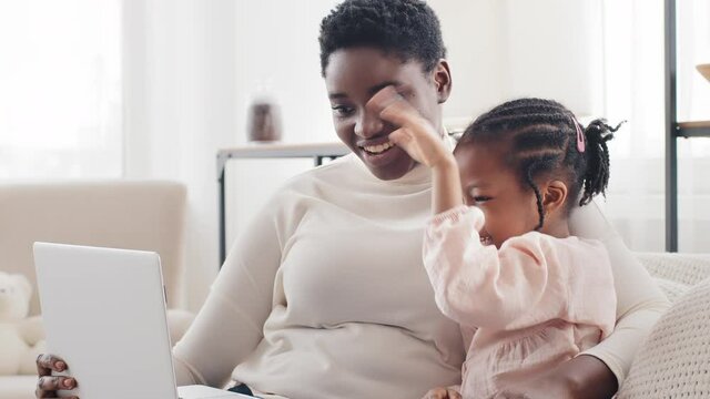 Portrait african afro american black ethnic mother sitting on sofa at home indoor with cute little daughter baby kid child waving goodbye looking at laptop online video chat conference watching movie