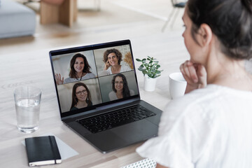 Fototapeta na wymiar back of a remote online coaching woman sitting on her work desk infront of laptop screen at her home office joining a team meeting or women circle of 4, coaching event or watching video conference