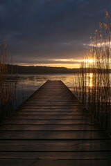 An old wooden pier on the shore of a lake at dusk in the golden light. Clouds in the sky and reeds in the water - image