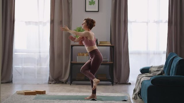 home fitness training of adult woman, lady is performing garudasana at living room of modern apartment