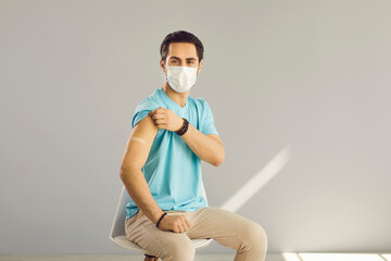 Young man with a medical face mask and a band-aid on his shoulder sits on a chair on a gray...
