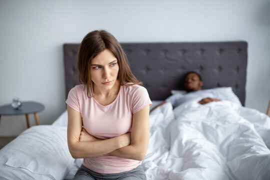 Depressed young woman having relationship problem, sitting on bed while her husband sleeping at home, copy space