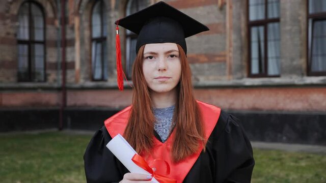 Portrait of a beautiful ginger female student dressed in a mantle and caps with a master's degree in hand university graduate on the background of a high school academy graduation day.