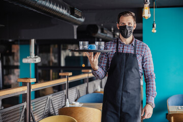 Serving coffee and water during the corona virus. Portrait of a male waiter in a plaid shirt and apron with a protective face mask walking around a cafe carrying a tray of coffee and water