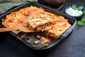 Traditional spiced Moroccan lamb ragout with filo pie served as close-up in a rustic black backing dish on a board