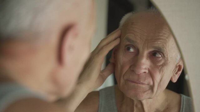 Gray haired man touching wrinkled face with hand looking in mirror