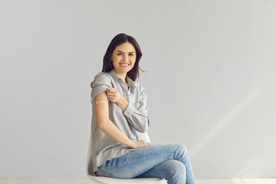 Woman promoting coronavirus vaccination and immunization campaign. Happy lady sitting on chair on gray studio background shows arm with adhesive patch after getting flu or Covid-19 vaccine injection