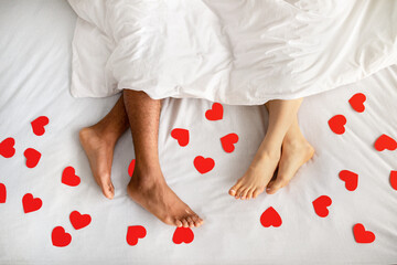 Top view of multiracial couple lying under blanket on bed with red paper hearts, closeup of feet