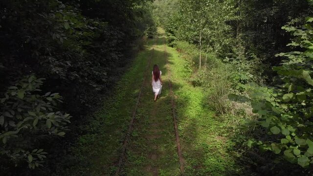 Aerial view of a young girl in a white dress from the back walking barefoot along an abandoned railway in a summer forest