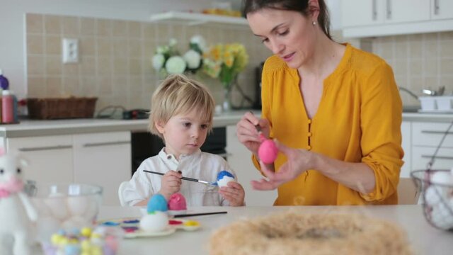 Beautiful blond child, toddler boy, painting easter eggs with mother at home, making easter wreath with rainbow colors