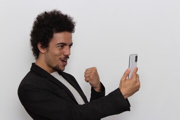 Man in black blazer on white background celebrating with cellphone in hand