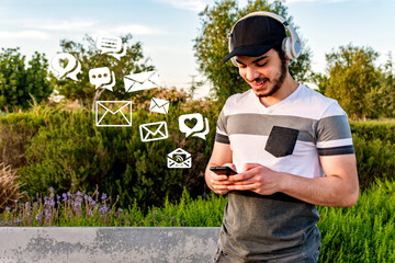 Young man with black cap and wireless headset sends messages with his smart phone in a park.