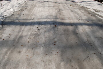 Snow-covered road with car tire tracks. It was a sunny spring day, a narrow road covered with snow on the snow there were imprinted footprints of people and tracks from car tires. Shadows from trees f