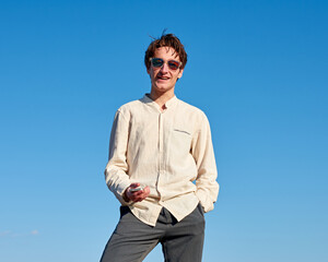 A Caucasian man holding his phone and smiling with one hand in pocket on sky background
