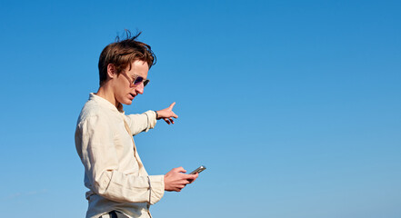 A Spanish white man with long hair, glasses and a beige shirt looking at his phone and pointing to his left on sky background