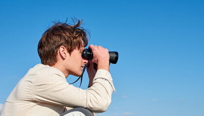 A Spanish white man in a beige shirt looking through binoculars on clear blue sky background