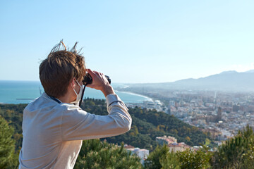 Fototapeta premium A close up of a young man from Spain in a white shirt wearing a mask observing the city of Málaga through binoculars