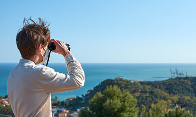 A shallow focus shot of a young man from Spain in a white shirt wearing a mask looking over the coastal town through binoculars
