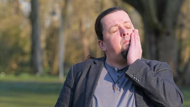 [4k] long haired goatee best ager is hat sweating in sunlight in public park