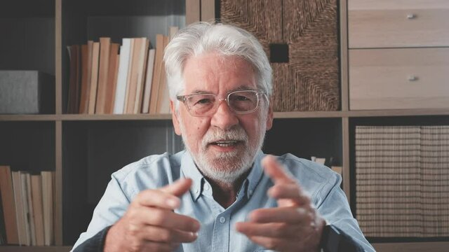 Head shot portrait confident old businessman coach wearing glasses looking at camera and talking, mentor speaker holding online lesson, explaining, sitting at wooden work desk in modern cabinet
