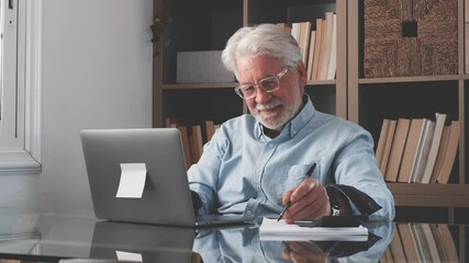 Happy old caucasian businessman smiling working online watching webinar podcast on laptop and learning education course conference calling make notes sit at work desk, elearning concept
