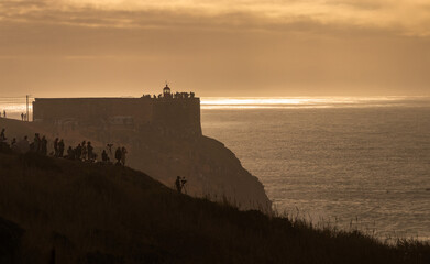 Fototapeta premium Fort next to the North beach in Nazaré, Portugal, seen in silhouette with people in the foreground watching the giant waves and in the background the sunlight mirrored in the sea water.