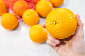 Organic fresh orange in hand, quality selection from red pattern net bag as blurred background. Tropical juicy refreshment fruit, full of vitamin c and fiber. Natural sweet and tasty. Depth of field.
