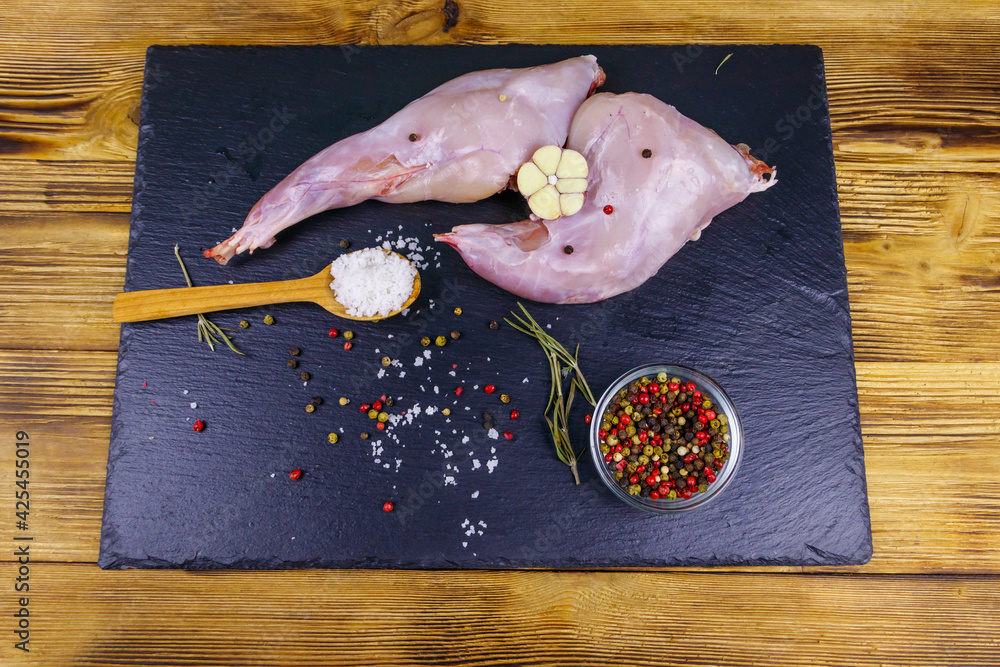 Wall mural Raw rabbit legs with spices on a black slate cutting board on wooden table. Top view