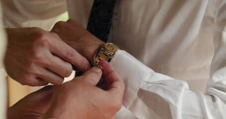 Groom getting ready for a wedding and having a groomsman assisting him with putting on a gold bracelet on his wrist. 