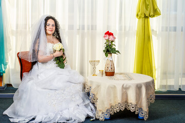 Fototapeta na wymiar A happy Jewish bride with a bouquet of white roses sits in the synagogue before the Huppa ceremony at a table with flowers and a menorah.