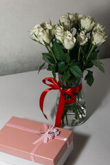 White tea roses stand in a glass vase, which is tied with a red ribbon and stands on the table. There is a gift nearby. in a pink box. Gift bouquet