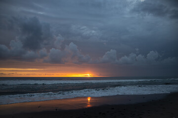 Incredibly beautiful sunset by the ocean in Bali. The bright sun sets in the water. The rays of the sun peeping out from behind the clouds and a beautiful reflection of the sunset in the water