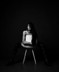 Obraz na płótnie Canvas Black and white. Excited gorgeous naked woman in high leather boots with laces sits backwards on white chair, hugging it over dark background. Fashion, vogue, sexy stylish look for woman concept