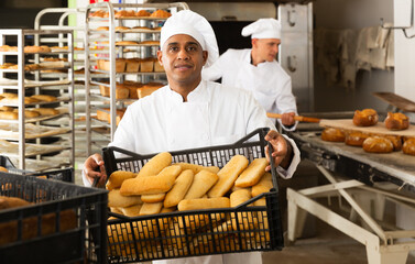 Positive man baker carrying tray with fresh baked bread at bakery kitchen