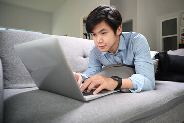 Happy young man lying on sofa in living room and surfing internet with computer laptop.
