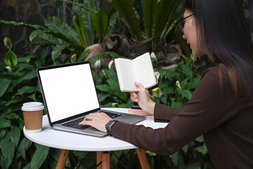 Side view young woman sitting at outdoor cafe holding notebook and working on laptop computer.