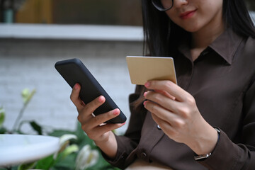 Cropped shot of young female holding credit card and using smart phone for paying bills online or shopping online.