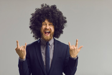 Office worker gone crazy. Funny excited cheerful handsome young man in suit, tie and Afro hair style wig, with happy face expression doing horn sign rock-n-roll Let's Party gesture on gray background