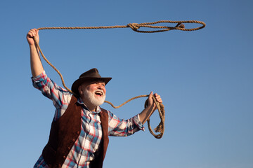 Old cowboy with lasso rope at ranch or rodeo. Bearded western man with brown jacket and hat catching horse or cow.