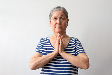 Elderly woman, gray-haired lady of the 60s in a striped white and blue dress praying with hands together asking for forgiveness smiling confident on a white background.